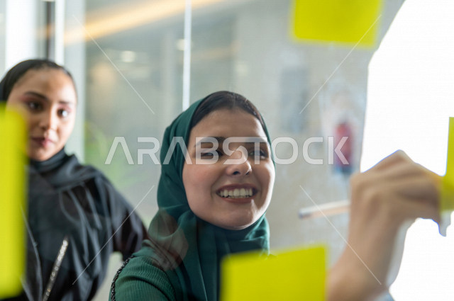 صورة مقربة لسيدة اعمال سعودية خليجية عربية مبتسمة ، اجتماع لسيدات اعمال سويا داخل الشركة ، شركة سعودية ، وظيفة مكتبية ، عمل خليجي ،  فريق العمل ، ورق ملاحظات ، خلف الزجاج