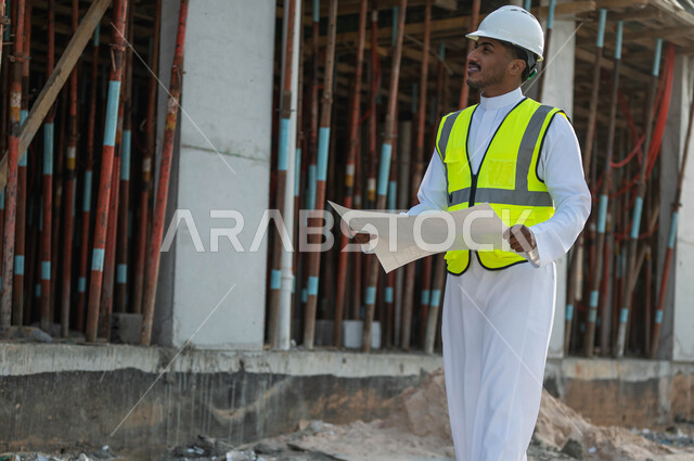 الاهتمام بالعمران والحرص على بناء الأساسيات الجيده للمبنى, العمل في القطاع الهندسي, مهندس عربي خليجي سعودي يرتدي الخوذة وسترة الحماية يحمل بيده مخطط التنظيم, النظر إلى شيء ما والتحقق من تطبيق المشاريع والخطط الهندسيه المعمارية في البناء
