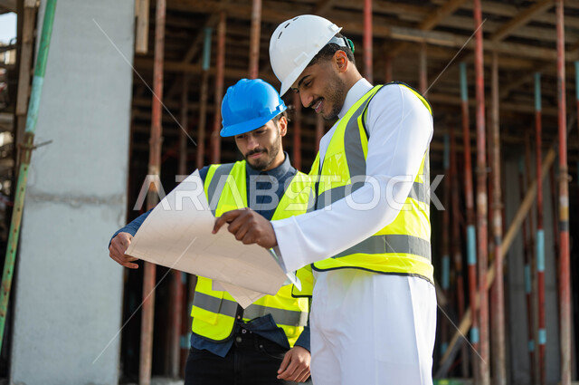 Consultations on completing work and proposing new ideas, supervising the implementation of projects and architectural plans in construction, two Saudi Gulf Arab workers and architects wearing helmets and protection vests holding the project plan, working in the engineering sector