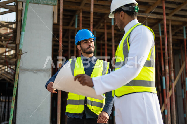 Proposing new ideas and consultations in completing the work, supervising the implementation of projects and architectural plans in construction, two Saudi Gulf Arab workers and architects wearing helmets and protection vests holding the project plan, working in the engineering sector.
