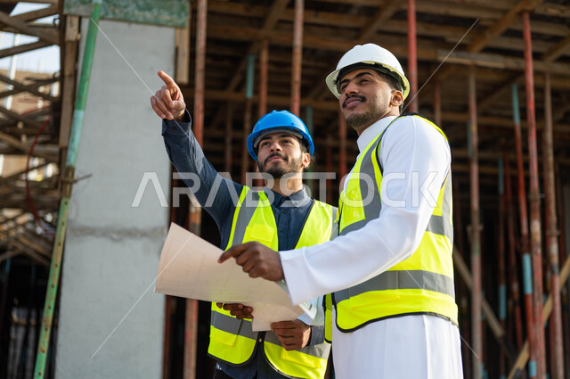 Verifying the application of the correct construction conditions, pointing and looking at something, two Saudi and Gulf Arab engineers wearing jackets and protection helmets standing at the work site, paying attention to the good foundations of the building, following up on the progress of the project’s organizational plan, discussion, dialogue and exchange of ideas.