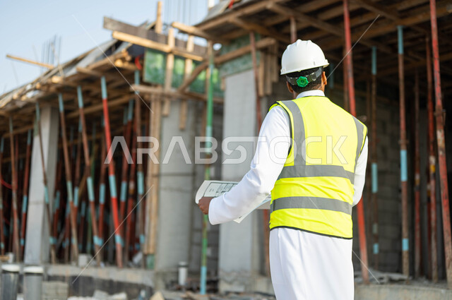المخططات والرسومات الإنشائيه, الطموح في العمل لتحقيق النجاح, صورة من الخلف لمهندس عربي سعودي خليجي يرتدي ستره الحمايه والخوذه ينظر إلى البناء, الاهتمام في جوده المواد الأوليه, التحقق من تطبيق المشاريع والخطط الهندسيه