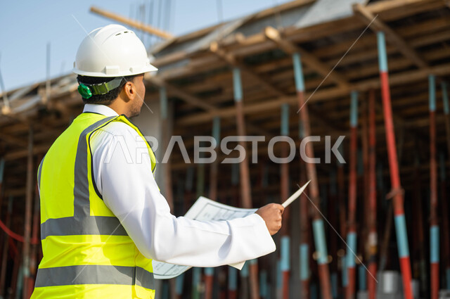 النظر نحو الأعلى و الدقه والمهاره في إتقان العمل , مهن ووظائف سعوديه, صورة من الخلف لمهندس عربي خليجي سعودي يرتدي الستره وخوذه الحمايه يحمل بيده مخطط الرسومات والإنشاءات الهندسيه , معاينة تطبيق الخطط والمشاريع الأساسية للمباني , إيماءات التركيز والاهتمام .