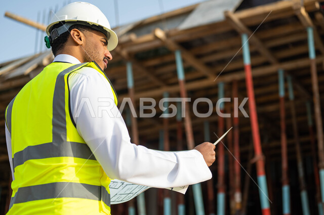 معاينة تطبيق الخطط والمشاريع الأساسية للمباني , مهن ووظائف سعوديه, الدقه والمهاره في إتقان العمل , صورة مقربة من الخلف لمهندس عربي خليجي سعودي يرتدي الستره وخوذه الحمايه ينظر إلى مخطط الرسومات والإنشاءات الهندسيه ,إيماءات التركيز والاهتمام .