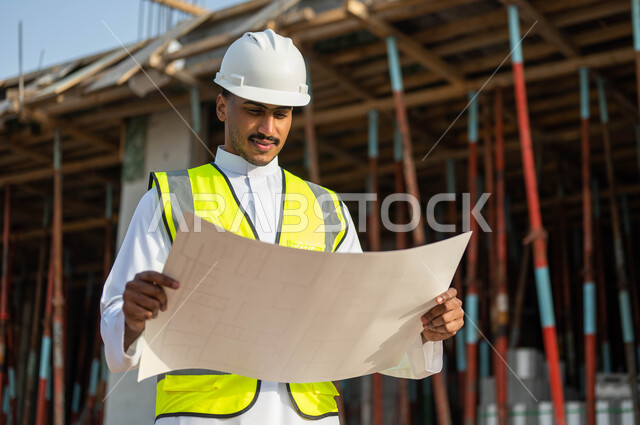 إيماءات التركيز والاهتمام , الدقه والمهاره في إتقان العمل , مهن ووظائف سعوديه,  مهندس عربي خليجي سعودي يرتدي الستره وخوذه الحمايه ينظر إلى مخطط الرسومات والإنشاءات الهندسيه , معاينة تطبيق الخطط والمشاريع الأساسية للمباني .