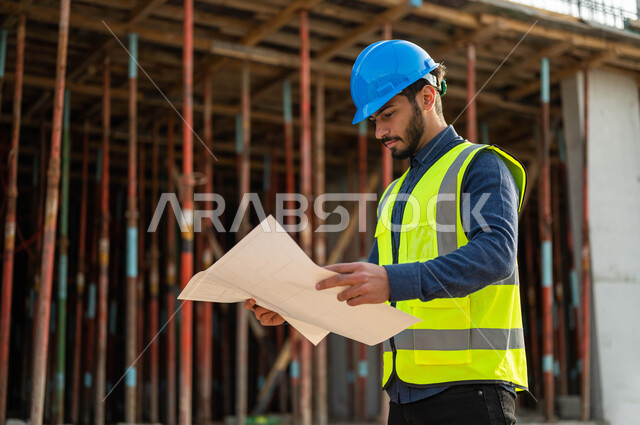 معاينة الأساسيات الجيده للمباني , صورة مقربة لمهندس عربي خليجي سعودي يرتدي الخوذة وسترة الحماية وينظر إلى مخطط المشروع بإيماءات التدقيق والاهتمام, العمل في القطاع الهندسي العمراني, التحقق من تطبيق المشاريع والخطط الهندسيه المعمارية.