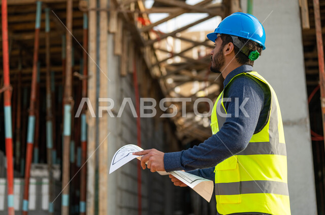 التحقق من تطبيق المشاريع والخطط الهندسيه المعمارية, صورة مقربة لمهندس عربي خليجي سعودي يرتدي الخوذة وسترة الحماية وينظر إلى البناء, العمل في القطاع الهندسي العمراني, الحرص على الأساسيات الجيده للمباني .