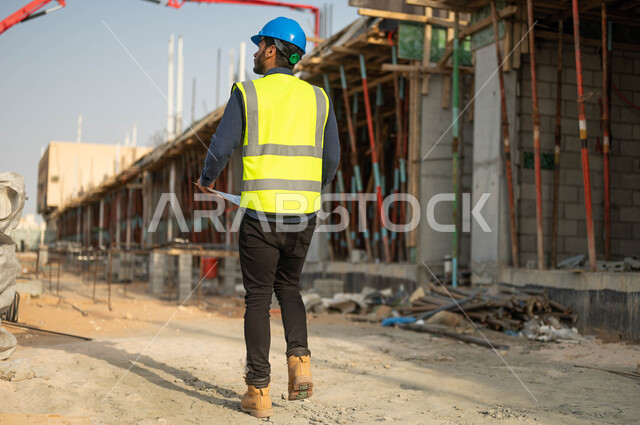 الفحص والتجول في مقر العمل ,  صورة من الخلف لمهندس عربي خليجي سعودي يرتدي الخوذة وسترة الحماية ويقوم بمقارنة المخطط الهندسي مع التنفيذ الواقعي للأعمال, التحقق من تطبيق المشاريع والخطط الهندسيه المعمارية, صورة الجسم بالطول الكامل .