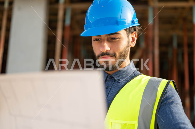 التحقق من تطبيق المشاريع والخطط الهندسيه المعمارية,  صورة مقربة لمهندس عربي خليجي سعودي يرتدي الخوذة وسترة الحماية وينظر إلى مخطط المشروع بإيماءات التدقيق والاهتمام,  الإنشاءات الاعمارية في الاراضي السعودية .