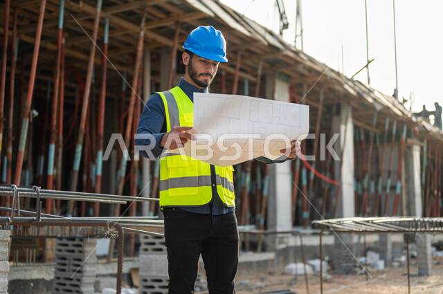 النظر إلى مخطط المشروع بإيماءات التدقيق والاهتمام, صورة مقربة لمهندس عربي خليجي سعودي يرتدي الخوذة وسترة الحماية ويقف أمام البناء, التحقق من تطبيق المشاريع والخطط الهندسيه المعمارية, الحرص على الأساسيات الجيده للمباني