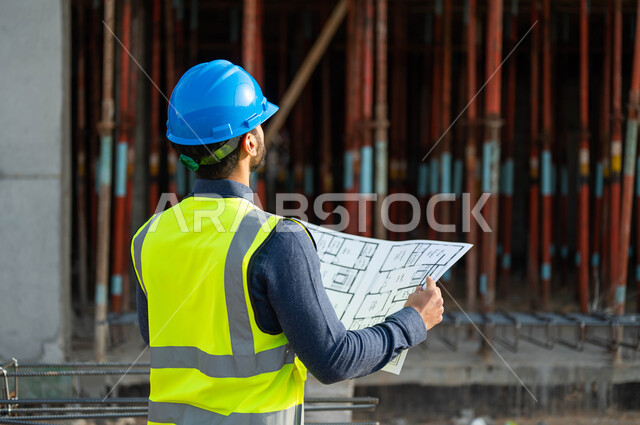 النظر نحو الأعلى و التحقق من تطبيق المشاريع الهندسيه المعمارية, صورة مقربة من الخلف لمهندس عربي خليجي سعودي يرتدي الخوذة وسترة الحماية ويقوم بمقارنة المخطط الهندسي مع التنفيذ الواقعي للأعمال ,  الحرص على وضع الأساسيات الجيده للمباني .