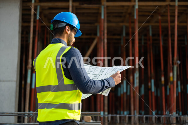 مفهوم الهندسة والانشاءات، الاشراف على تطبيق المشاريع و الخطط الهندسيه في البناء ،الحرص على بناء الأساسيات الجيده للمباني،صورة من الخلف  لمهندس عربي خليجي سعودي يرتدي الخوذة وسترة الحماية يحمل بيده المخطط الهندسي ويقوم بدراسته وتفحصه، مهن ووظائف سعودية
