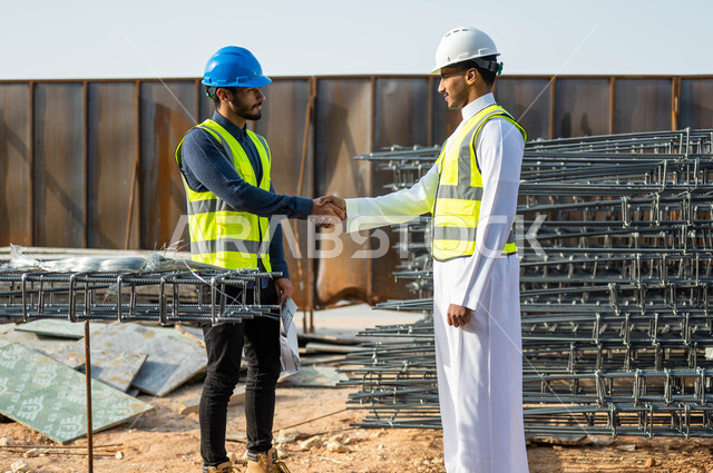 Handshake, welcome and reception gestures, the concept of cooperation and joint work, two Saudi Gulf Arab workers and civil engineers wearing helmets and protection vests shake hands and agree on engineering plans and projects at the work site, protected construction in Saudi territory