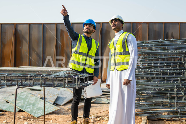 The concept of cooperation and joint work, pointing to something, conversations and chats revolving around the project, two Saudi Gulf Arab workers and engineers wearing helmets and protection vests supervising the implementation of engineering projects in Saudi Arabia, comparing the engineering plan with work on the ground