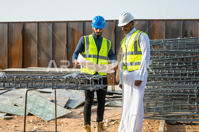 Following up on the progress of construction projects, paying attention to building good foundations for buildings, verifying the correctness of the application of architectural engineering plans, two Saudi Gulf Arab engineers wearing helmets and protection vests exchanging ideas and opinions to develop engineering work