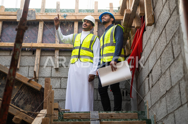 التأكد من وضع الأساسيات الجيده في المباني , الأحاديث والدردشات التي تدور حول المشروع، عامل ومهندس عربيان خليجيان سعوديان يرتديان الخوذة وسترة الحماية يعملان في مجال الهندسة المعمارية ،الاشراف على تطبيق الخطط و المشاريع الهندسيه في السعودية  .