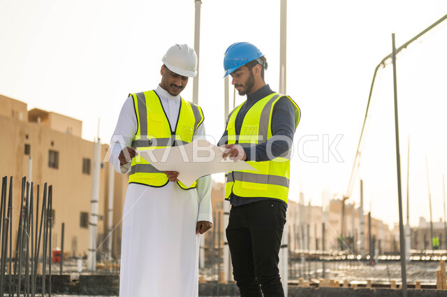 مفهوم العمل المشترك ، الاهتمام في بناء الأساسيات الجيده للمباني، متابعة سير المشاريع الإعمارية والتحقق من صحه تطبيق الخطط الهندسيه , مهندسان عربيان خليجيان سعوديان يرتديان الخوذة وسترة الحماية يتبادلان الأفكار والآراء لتطوير العمل .