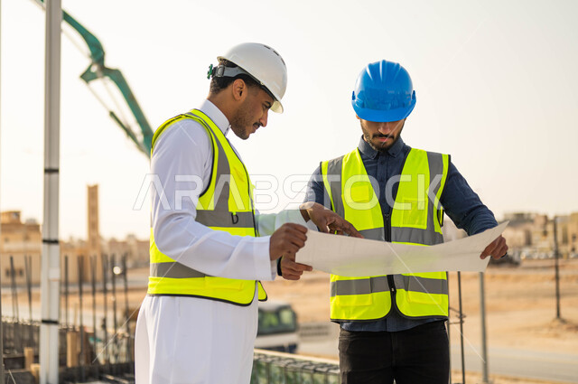 Following up on the progress of construction projects and comparing the engineering plan with the work on the ground, exchanging ideas and discussions about the work project, interest in building good basics for buildings, two Saudi Gulf Arab engineers wearing helmets and protection vests discussing work plans