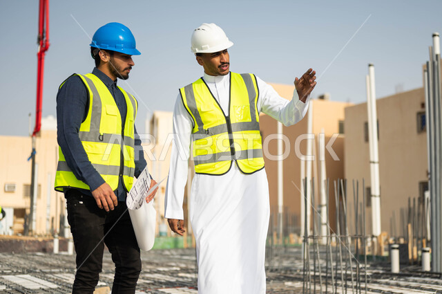 The concept of joint work, following up on the progress of construction projects and verifying the implementation of engineering plans in buildings, two Saudi Gulf Arab engineers wearing helmets and protection vests exchanging ideas, looking at something and pointing to something