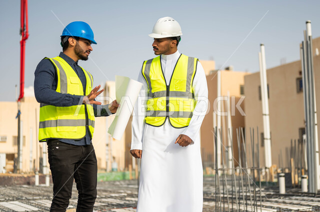 التشارك في الأعمال الهندسية ، الاهتمام في بناء الأساسيات الجيده للمبانى، متابعة سير المشاريع الإعمارية, مفهوم العمل المشترك ,مهندسان عربيان خليجيان سعوديان يرتديان الخوذة وسترة الحماية يقفان في مقر العمل