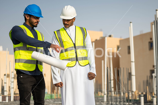 Construction construction in Saudi territory, gestures of looking and pointing at something, verifying the correctness of the application of architectural engineering plans in buildings, two Saudi Gulf Arab engineers wearing helmets and protection vests exchanging ideas and discussions about the work project