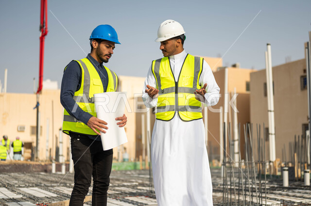 Discussions and follow-up of the progress of construction projects, the concept of cooperation and joint work, verifying the application of engineering plans in buildings, two Saudi Gulf Arab engineers wearing helmets and protection vests and exchanging opinions, different facial and hand gestures