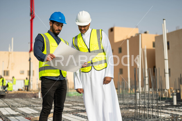 مفهوم التعاون والعمل المشترك , تبادل الأفكار والنقاشات حول المشاريع الهندسية ، مهندسان عربيان خليجيان سعوديان يرتديان الخوذة وسترة الحماية يدققان مخطط البناء, الاهتمام في بناء الأساسيات الجيده للمباني، متابعة سير المشاريع الإعمارية  .