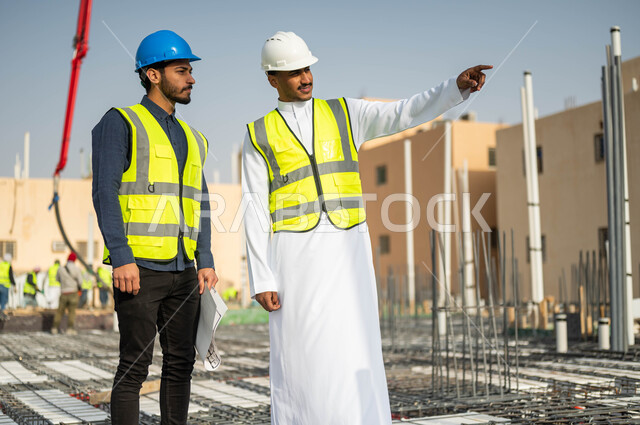 متابعة سير المشاريع الإعمارية والتحقق من تطبيق الخطط الهندسية , الاشارة إلى شيء ما ، الحرص على بناء الأساسيات الجيده للمباني، عامل ومهندس عربيان خليجيان سعوديان يرتديان الخوذة وسترة الحماية يتبادلان الأفكار الهندسية في موقع العمل.