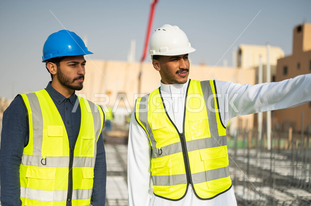 الاشارة إلى شيء ما ،متابعة سير المشاريع الإعمارية و التحقق من تطبيق الخطط الهندسيه ، الحرص على بناء الأساسيات الجيده  للمباني، عامل ومهندس عربيان خليجيان سعوديان يرتديان الخوذة وسترة الحماية في موقع العمل .