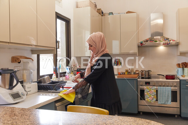 استخدام ملمع زجاج ورخام فعال، مفهوم النظافة والعناية بترتيب المنزل، امرأة عربيه خليجية اماراتية محجبه ترتدي عباءة سوداء تقوم بتنظيف المطبخ، الاهتمام بقواعد الوقاية والحماية من الجراثيم والميكروبات.