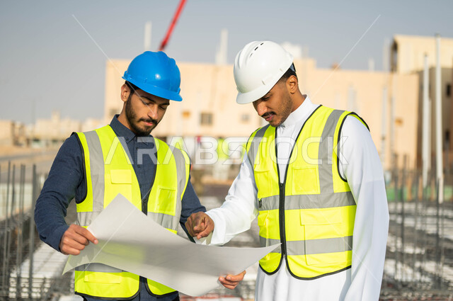 المناقشة والتشاور في اقتراح أفكار هندسية جديدة ، مهندسان مدنيان عربيان خليجيان سعوديان يرتديان الخوذة وسترة الحماية يمسكان المخطط ويتبادلان الافكار ، الأحاديث والدردشات التي تدور حول المشروع، الاشراف على تطبيق الخطط و المشاريع المعمارية في السعودية