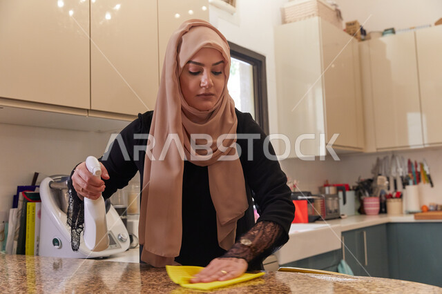 The concept of cleanliness and care for house arrangement, a veiled Arab Gulf Emirati woman wearing a black cloak cleans the kitchen, paying attention to the rules of prevention and protection from germs and microbes, using an effective glass and marble p