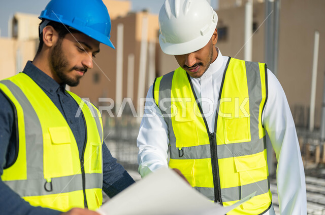 الاشراف على تطبيق الخطط و المشاريع الهندسيه في السعودية ،مهندسان مدنيان عربيان خليجيان سعوديان يرتديان الخوذة وسترة الحماية يمسكان مخطط البناء ويتبادلان الأفكار الهندسية، الأحاديث والدردشات التي تدور حول المشروع بإيماءات السرور .