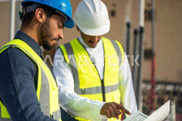 الأحاديث والدردشات التي تدور حول المشروع بإيماءات السرور، المساعدة في اقتراح أفكار جديدة ,مهندسان معماريان عربيان خليجيان سعوديان يرتديان الخوذة وسترة الحماية يقفان في موقع العمل ، الاشراف على تطبيق الخطط و المشاريع الهندسيه في السعودية .