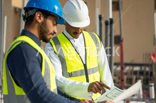 المساعدة في اقتراح أفكار هندسية جديدة ،مهندسان مدنيان عربيان خليجيان سعوديان يرتديان الخوذة وسترة الحماية يمسكان المخططات الهندسية في موقع العمل ، الأحاديث والدردشات التي تدور حول المشروع بإيماءات السرور، تبادل الأفكار والآراء حول المشروع