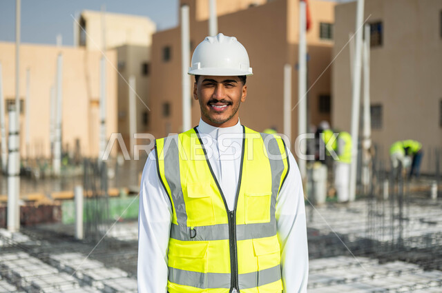 الخطط و المشاريع الهندسيه المعمارية في موقع العمل ، صورة مقربة لمقاول عربي خليجي سعودي يقف باستقامة و يرتدي الخوذة وسترة الحماية ينظر إلى الكاميرا، العمل في القطاع الهندسي , النمو والتطور العمراني على أيدي أبناء الوطن.