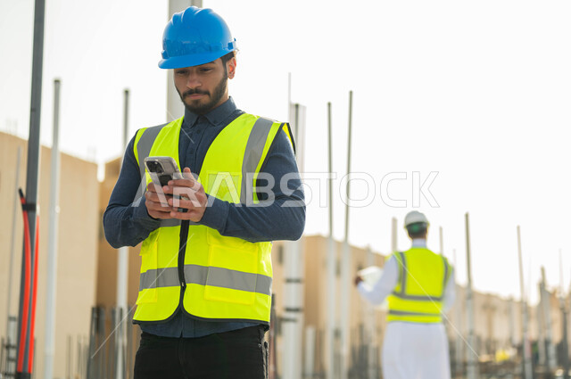 دمج العمل مع التقنية و التكنولوجيا ، الاطلاع على تصاميم هندسية جديدة عبر الجوال، مهندس مدني عربي خليجي سعودي يرتدي الخوذة وسترة الحماية يستخدم الهاتف المحمول في موقع العمل ،النمو والتطور العمراني في السعودية , مجال الانشاءات والمشاريع الهندسية المعمارية