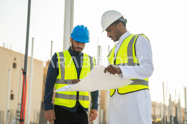 Help in proposing new ideas, supervising the implementation of engineering plans and projects in Saudi Arabia, two Saudi Gulf Arab civil engineers wearing helmets and protection vests carrying plans, conversations and chats revolving around the project