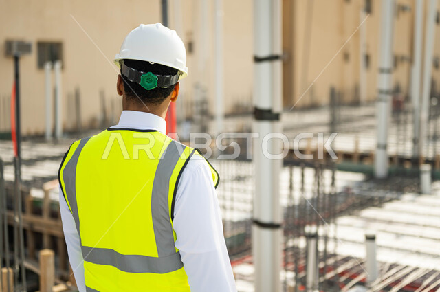 تطور وتقدم المباني على يد مهندسين سعوديين, بناء الاساسيات الجيدة , الاهتمام في صحه تطبيق الخطط الهندسيه المعمارية في المبنى ,صورة مقربة من الخلف لمهندس عربي خليجي سعودي يرتدي الخوذة وسترة الحماية يقف في موقع التنفيذ ، مهن ووظائف سعودية