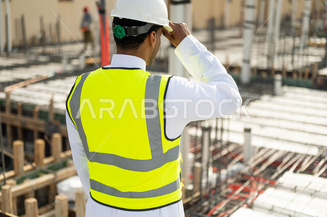 Progress and development of buildings at the hands of Saudi engineers, building good foundations, paying attention to the correct application of architectural plans in the building, a close-up photo from the back of a Saudi Gulf Arab engineer wearing a helmet and a protective vest standing at the implementation site, Saudi professions and jobs