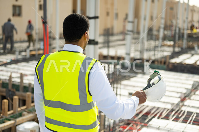 تطور وتقدم المباني على يد مهندسين سعوديين, بناء الاساسيات الجيدة , الاهتمام في صحه تطبيق الخطط الهندسيه المعمارية في المبنى ,صورة مقربة من الخلف لمهندس عربي خليجي سعودي يقوم بارتداء الخوذة و يقف في موقع التنفيذ ، مهن ووظائف سعودية