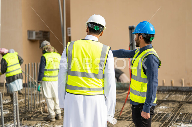 بناء الاساسيات الجيدة للمبنى, الاهتمام في صحه تطبيق الخطط الهندسيه المعمارية في المبنى ,صورة مقربة من الخلف لمهندسان عربيان خليجيان سعوديان يرتديان الخوذة وسترة الحماية يشرفان على مجموعة من العمال في موقع العمل والاشارة إلى شيء ما