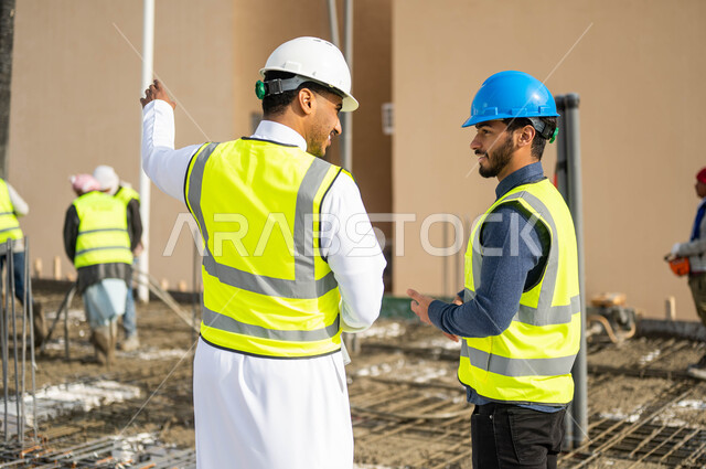 متابعة سير المشاريع الإعمارية, الاهتمام في صحه تطبيق الخطط الهندسيه المعمارية في المبنى ,صورة مقربة من الخلف لمهندسان عربيان خليجيان سعوديان يرتديان الخوذة وسترة الحماية يشرفان على مجموعة من العمال في موقع العمل والاشارة إلى شيء ما