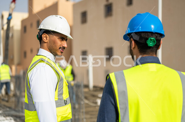 بناء الأساسيات الجيده للمبنى، متابعة سير المشاريع الإعمارية, الاهتمام في صحه تطبيق الخطط الهندسيه المعمارية في المبنى ,صورة مقربة من الخلف لمهندسان عربيان خليجيان سعوديان يرتديان الخوذة وسترة الحماية يشرفان على مجموعة من العمال في موقع العمل