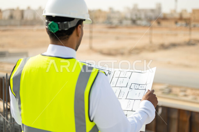 الوقوف في موقع تنفيذ المشروع ,الرقابة على المشاريع الهندسية و تدقيق الاعمال, مهن ووظائف سعودية، تصاميم معمارية حديثة, وظائف و مهن سعودية، صورة مقربة من الخلف لمهندس معماري عربي خليجي سعودي يرتدي الخوذة وسترة الحماية يعمل في القطاع الهندسي