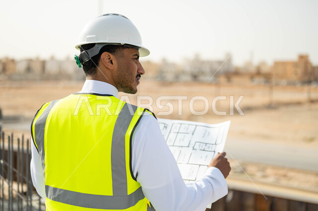 الرقابة على المشاريع الهندسية و تدقيق الاعمال, مهن ووظائف سعودية، تصاميم معمارية حديثة, العمل في القطاع الهندسي ،وظائف و مهن سعودية، صورة مقربة من الخلف لمهندس معماري عربي خليجي سعودي يرتدي الخوذة وسترة الحماية يقف في موقع تنفيذ المشروع