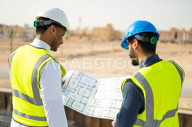 تصاميم معمارية حديثة, تبادل الاراء والافكار حول الرسومات والمخططات الانشائية، الاهتمام في بناء الأساسيات الجيده، متابعة سير المشاريع الإعمارية, صورة مقربة من الخلف لمهندسان عربيان خليجيان سعوديان يرتديان اللباس الخاص بالعمل يمسكان المخطط الهندسي