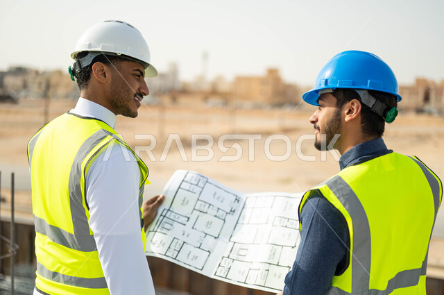 تصاميم معمارية حديثة, تبادل الاراء والافكار حول الرسومات والمخططات الانشائية، الاهتمام في بناء الأساسيات الجيده، متابعة سير المشاريع الإعمارية, صورة مقربة من الخلف لمهندسان عربيان خليجيان سعوديان يرتديان اللباس الخاص بالعمل يمسكان المخطط الهندسي