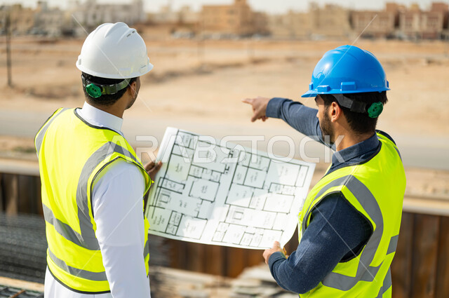 Referring to the location of the project, drawings and construction plans, attention in building the good basics of buildings, following up the progress of reconstruction projects, a picture close to the back of an engineer and Arab Gulf Gulf, two Saudi Gulf workers, wearing the work dress that holds the engineering plan.