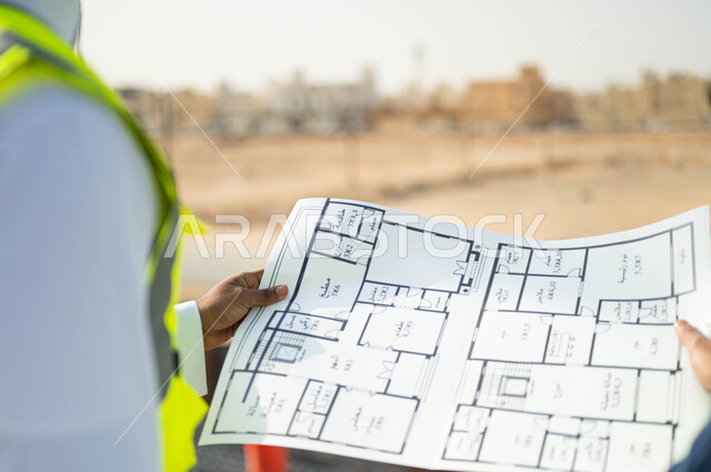 الرسومات والمخططات الانشائية، الاهتمام في بناء الأساسيات الجيده، متابعة سير المشاريع الإعمارية, التحقق من صحه تطبيق الخطط الهندسيه المعمارية أثناء إنشاء المبنى ,صورة مقربة من الخلف لمهندسان عربيان خليجيان سعوديان يقومان بمقارنة المخطط الهندسي مع العمل على ارض الواقع